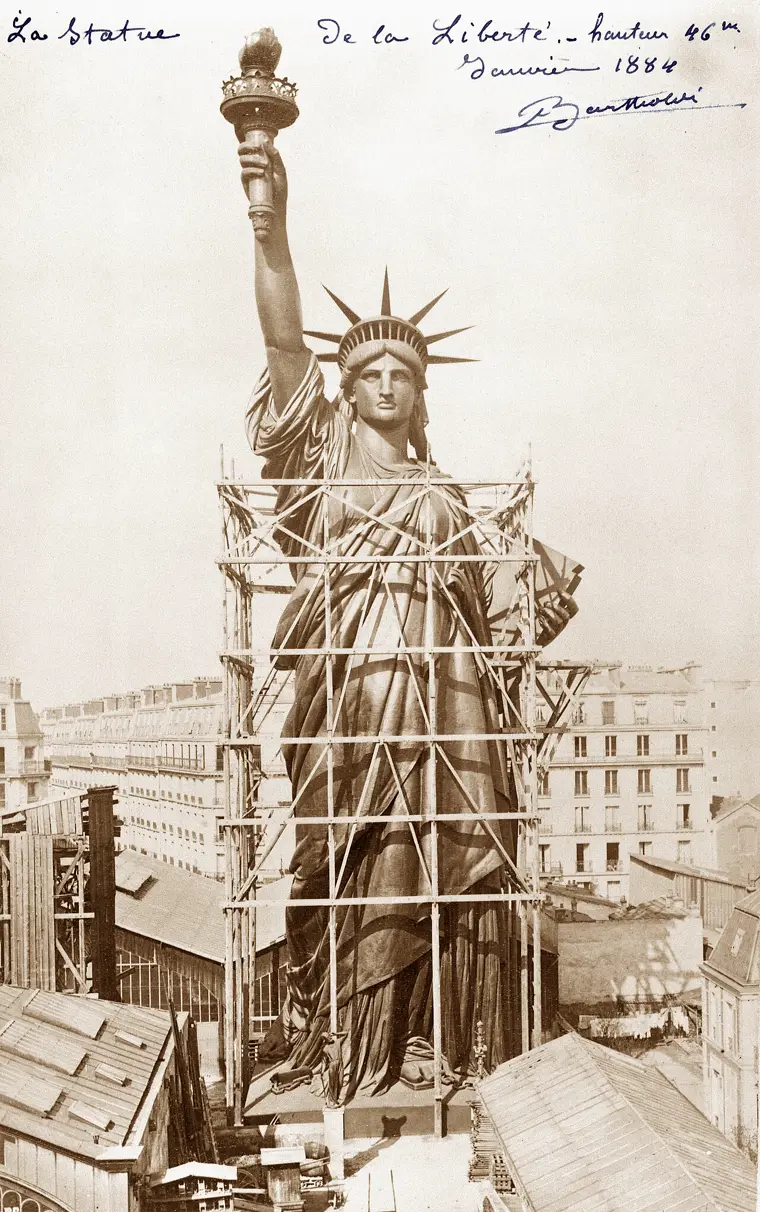 Black-and-white photograph from 1884: the Statue of Liberty, fully assembled and wrapped in scaffolding, towers over the rooftops of a Paris street. Bartholdi's handwritten note in the corner gives her height, 46 meters.